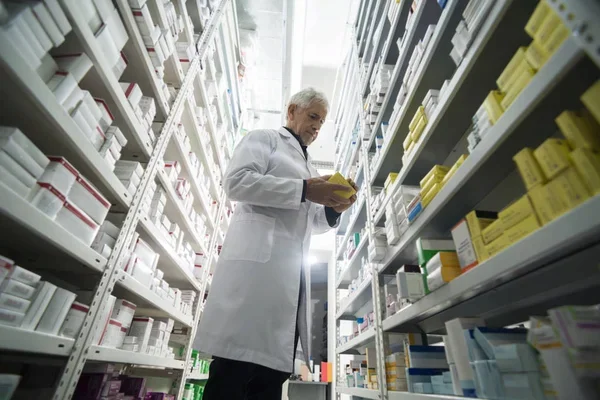 Farmacéutico mayor con bata blanca inspecciona una pequeña caja amarilla mientras está de pie en el pasillo de un almacén médico. Está rodeado de estanterías altas de metal repletas de numerosas cajas de medicamentos blancas y amarillas