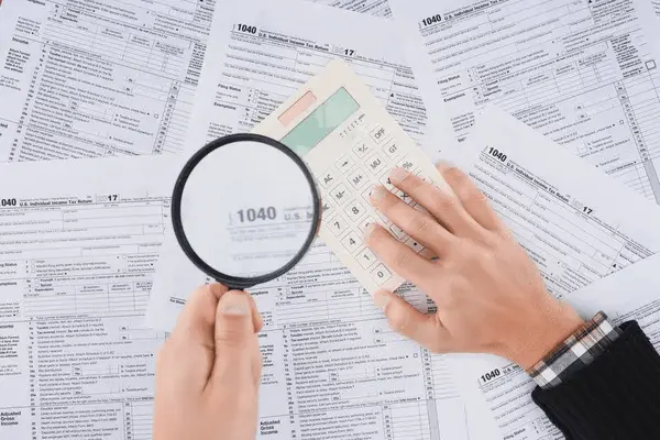 Vista recortada del hombre sosteniendo lupa y utilizando calculadora con formularios de impuesto a la renta empresarial sobre fondo
