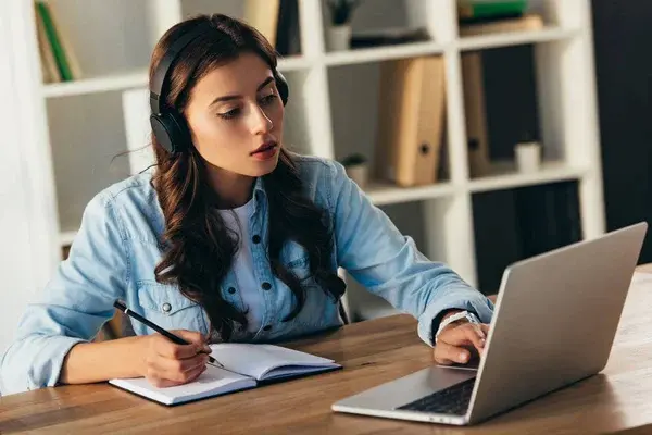 Estudiante joven en su escritorio con audífonos, tomando notas mientras atiende un curso en línea sobre el NRUS desde su laptop